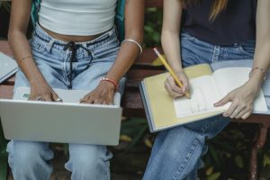High angle crop faceless multiracial female friends typing on laptop and drawing in notebook with pencil while sitting on bench
