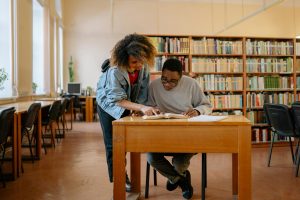 Two students engage in studying together at a library, surrounded by books and a peaceful atmosphere.