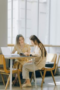 Two women collaborating over a laptop and book in a well-lit indoor setting.